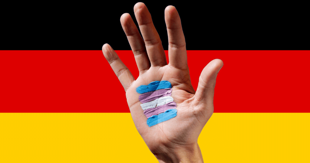 Bandera oficial de Alemania de fondo con una mano asomando que sostiene o muestra la bandera trans, representando la aprobación de la nueva ley de identidad.