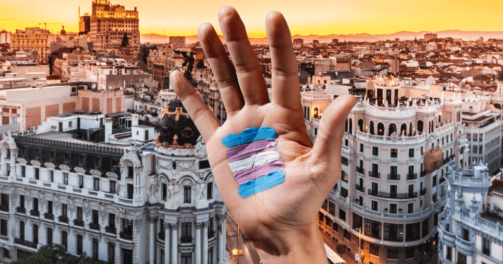 Vista aérea de un plano del centro de Madrid sobre el que asoma una mano con la bandera trans pintada, simbolizando la protección de derechos en la capital.