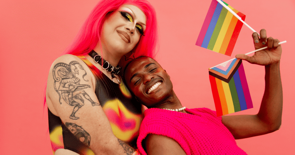 Mujer trans y hombre sonrientes, mirando a cámara. El hombre sostiene dos banderas LGTBI+.