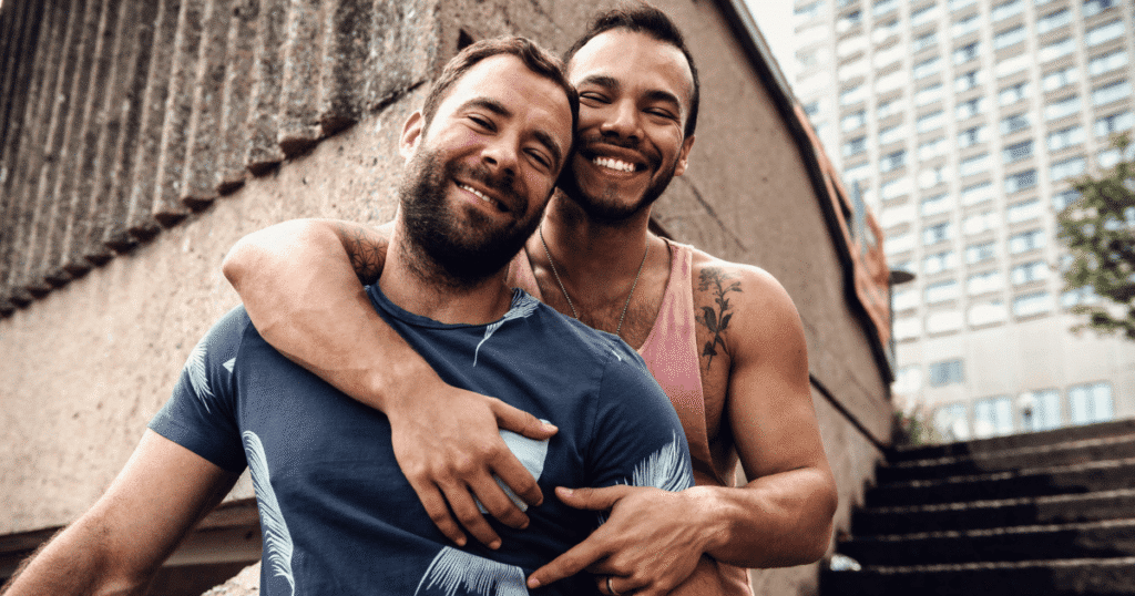 Primer plano de una pareja de hombres homosexuales, abrazados y sonriendo directamente a la cámara.