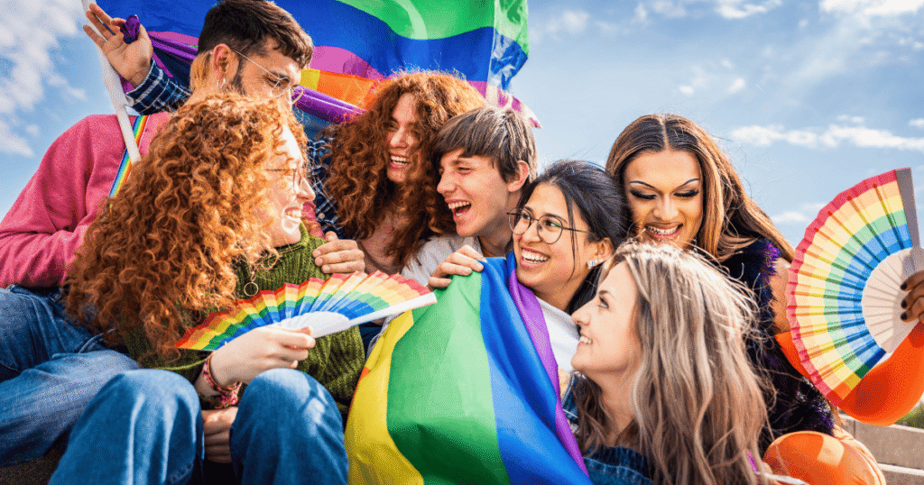 Grupo diverso de jóvenes sonriendo y mirándose, agitando abanicos y banderas LGTBI+, celebrando con alegría.