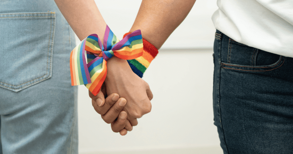 Primer plano de dos manos agarradas con pulseras y lazos de colores LGTBI+ y arcoíris, simbolizando la unidad y la resistencia contra la discriminación.