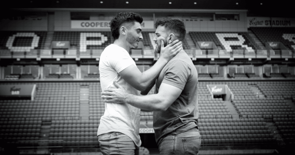 Josh Cavallo en el centro de un campo de fútbol sujetando cariñosamente a su marido del cuello para besarlo en una foto en blanco y negro.